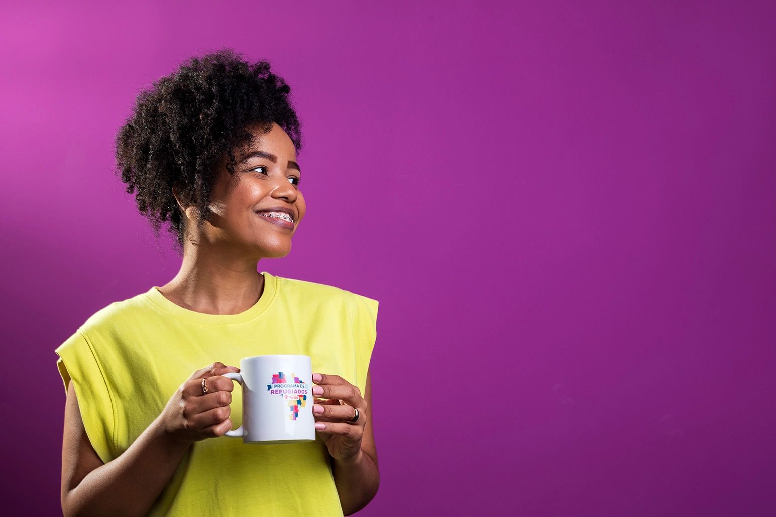 Fotografia institucional — colaboradora no escritório da Tenda — Colaboradora de camisa amarela em fundo roxo, segurando uma caneca, no escritório da Tenda Construtora. Uso institucional.